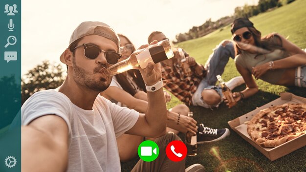 Auto-retrato de jovens sorridentes em trajes casuais, desfrutando de comidas e bebidas ao ar livre durante a videochamada