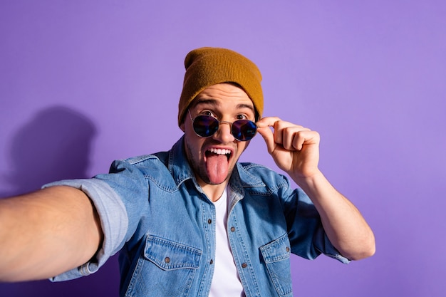 Auto-retrato de um homem tolo, casual engraçado, hilário, ridículo, tirando uma selfie mostrando a língua usando uma jaqueta jeans, boné marrom, chapéu isolado sobre fundo de cor roxa vívida