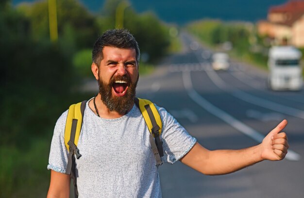 Foto auto-stop-reisen hipster versuchen, das auto zu stoppen und daumen nach oben geste mann mit fröhlichem gesicht und bart