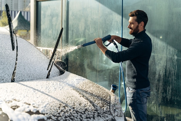 Auto waschen. Autowäsche an dem besonderen Ort allein. Mann lächelt in die Kamera, während er schwarzes Auto wäscht. Autoreinigung mit Hochdruckwasserkonzept