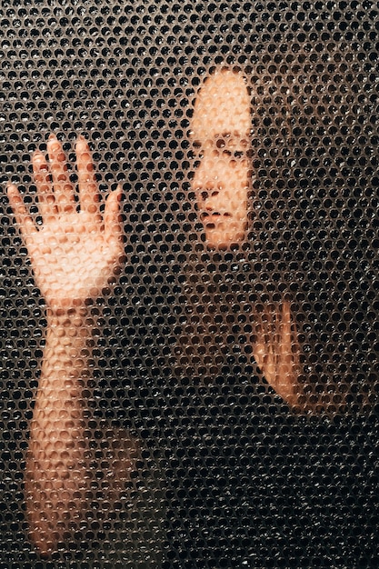 Autoaislamiento de Covid-19. Melancolía pandémica. Riesgo de infección. Retrato artístico texturizado de una mujer infeliz y cansada de negro tocando la pared de envoltura de burbujas de plástico sola en la oscuridad.