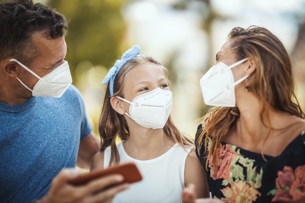 Autoaislamiento en la naturaleza. Joven familia feliz con máscara protectora N95 divirtiéndose durante la caminata en el parque y usando el teléfono inteligente.