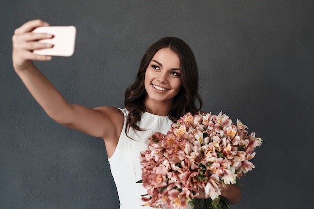 Autofoto perfecto. Atractiva mujer joven sonriente con ramo de flores tomando selfie mientras está de pie contra el fondo gris