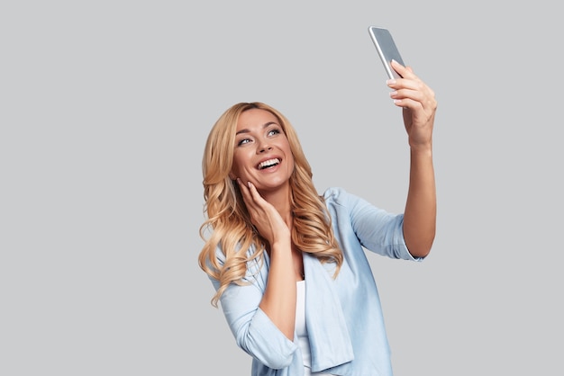 Autofoto perfecto. Atractiva mujer joven tomando selfie y sonriendo mientras está de pie contra el fondo gris