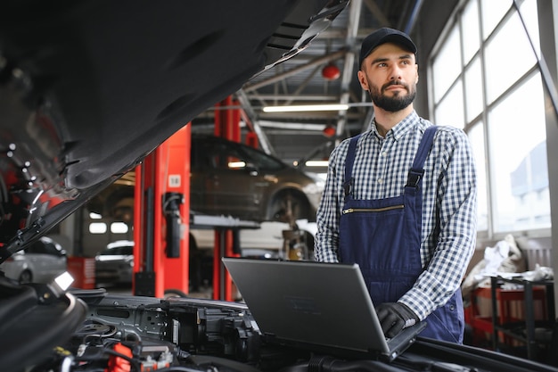 Un automecánico barbudo con uniforme de trabajo está de pie junto a un automóvil roto y usa la computadora portátil especial para el mantenimiento del automóvil