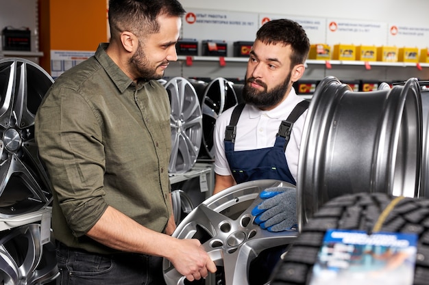 Automechaniker, der dem Kunden das beste Autorad anbietet. selbstbewusster bärtiger Mann im Uniformgespräch mit dem Kunden kam, um neue Räder zu kaufen