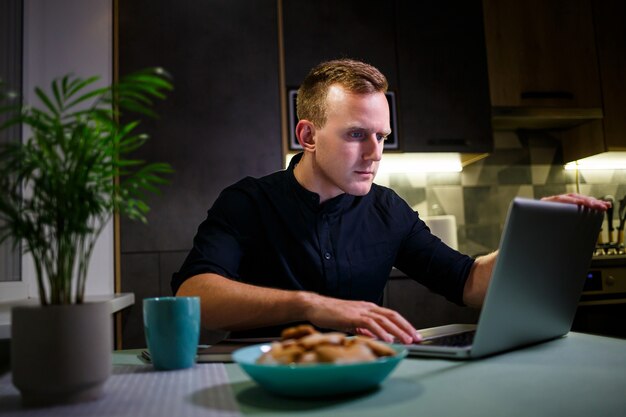 Un autónomo trabaja desde casa en la cocina y usando una computadora portátil. Hombre exitoso que trabaja con una computadora portátil y lee buenas noticias. Apuesto empresario exitoso se sienta y trabaja en su casa moderna.