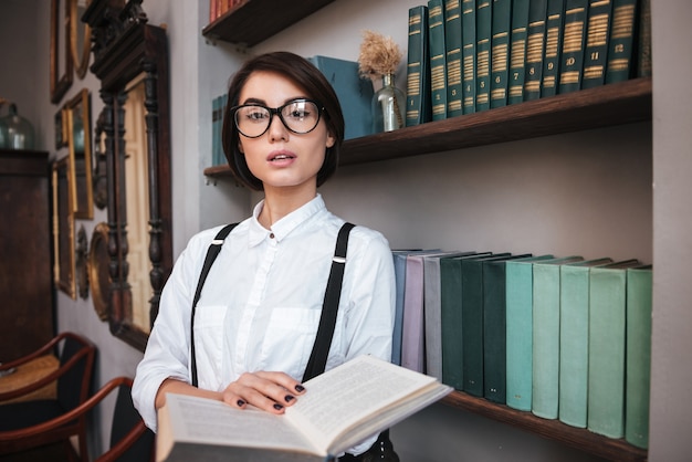 Autorin in Brille und weißem Hemd, die neben dem Bücherregal steht und ein Buch hält