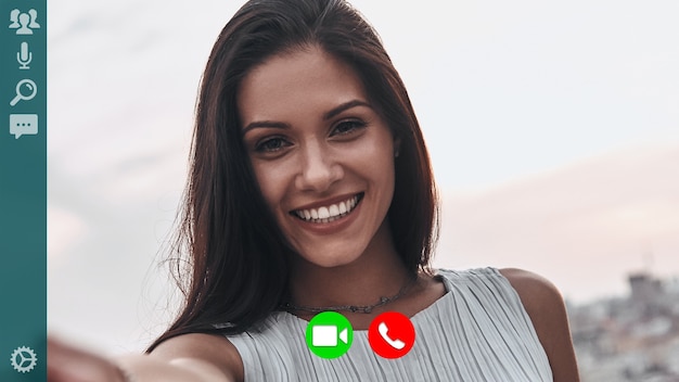 Foto autorretrato de mujer joven encantadora sonriendo y mirando a cámara mientras tiene videollamada