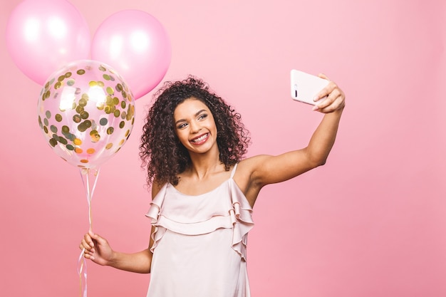 Autorretrato de niña afroamericana positiva en vestido con globos en la mano disparando selfie en cámara frontal aislada sobre fondo rosa.