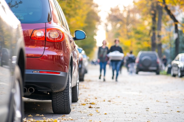 Autos, die an einem hellen Herbsttag in einer Reihe auf einer Straßenseite der Stadt geparkt sind, mit unscharfen Menschen, die auf der Fußgängerzone spazieren.