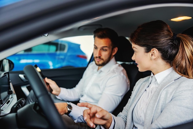 Autoverkäufer, der mit einem Kunden in einem Auto sitzt und ihm Vorteile und Leistungen eines Autos zeigt