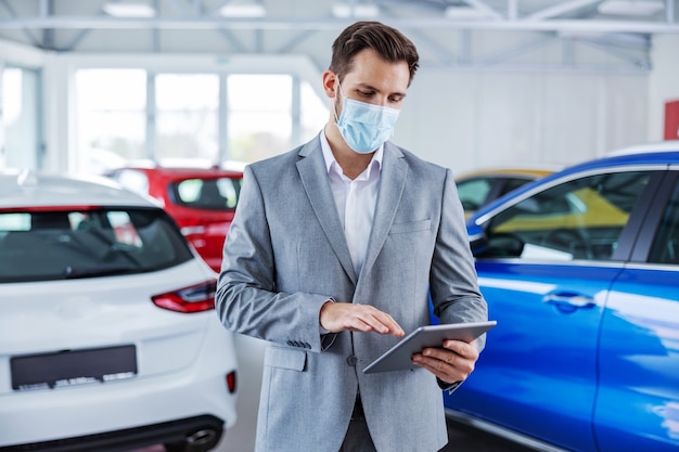 Autoverkäufer mit Gesichtsmaske beim Stehen im Autosalon und beim Verwenden des Tablets zur Online-Überprüfung des Lagers