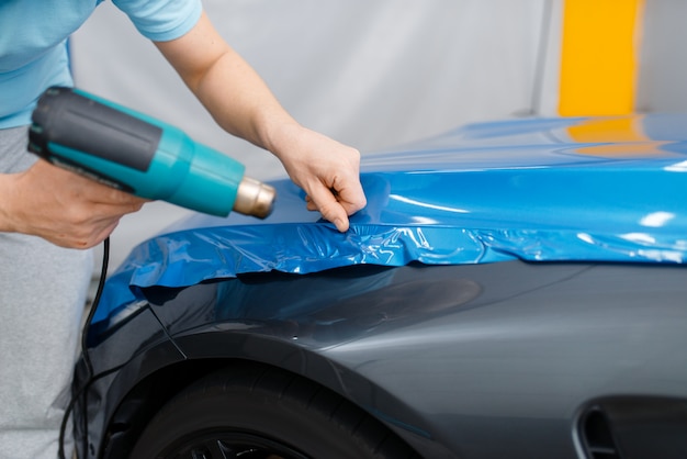 Autoverpackung, mechaniker mit trockner installiert schützende vinylfolie  oder folie auf der motorhaube. arbeiter macht automatische detaillierung.  autolackschutzbeschichtung, professionelles tuning