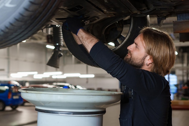 Autowartung Ein männlicher Automechaniker in Uniform wechselt das Öl im Motor, während er unter dem Auto auf der Hebebühne steht