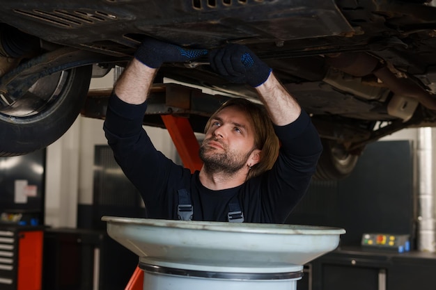 Autowartung Ein männlicher Automechaniker in Uniform wechselt das Öl im Motor, während er unter dem Auto auf der Hebebühne steht