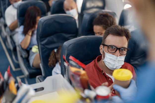 Auxiliar de vuelo que sirve bebidas a un pasajero masculino con mascarilla protectora a bordo. Viajar en avión durante la pandemia de Covid19. Enfoque selectivo