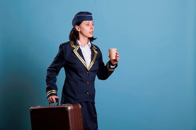 Auxiliar de vuelo con uniforme de aviación profesional llegando tarde, llevando maleta. Azafata con equipaje caminando en el aeropuerto, mujer sosteniendo una taza de café, plano medio de vista lateral