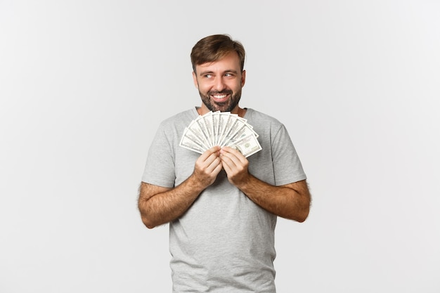 Avaricioso hombre codicioso sonriente con barba, pensando en ir de compras, sosteniendo dinero y mirando hacia arriba