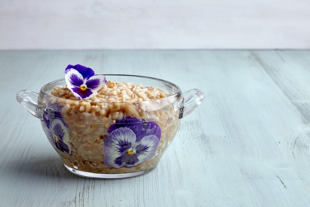 Aveia de grãos inteiros em uma tigela de vidro guarnecida com flores de viola em uma mesa de madeira azul