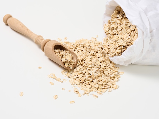 Avena cruda en una bolsa de papel blanco y una cuchara de madera sobre una mesa blanca, papilla de desayuno