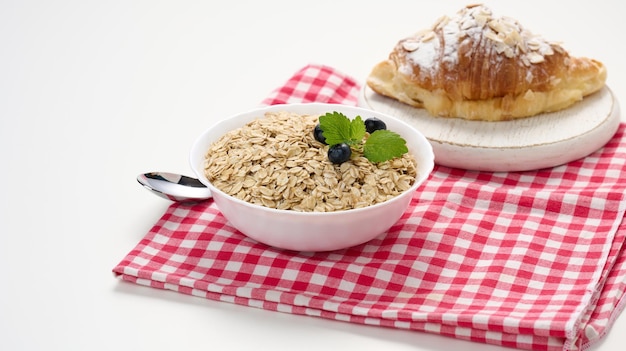 Avena cruda en plato de cerámica blanca y croissant en desayuno de mesa blanca