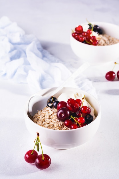 Avena cruda con yogur y bayas de cereza, grosella y arándano para hacer un desayuno saludable en un tazón de cerámica blanca sobre una luz