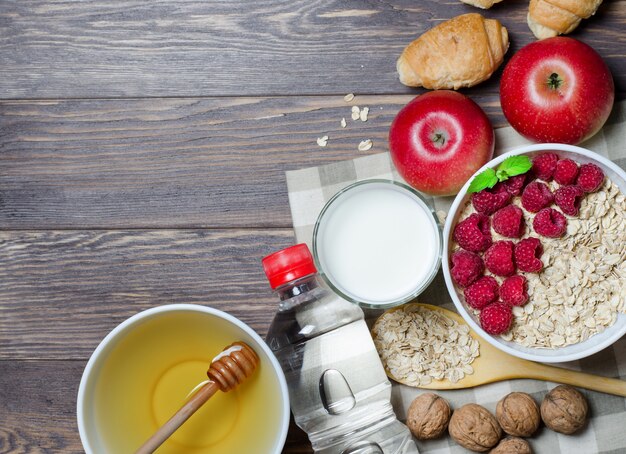 Avena con frambuesa, leche en un vaso, miel, agua mineral en una botella, nuez, croissant, manzanas rojas.