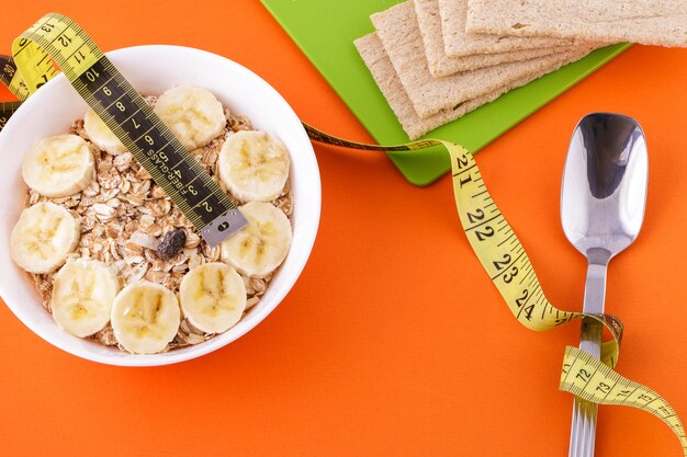 Avena con plátano en rodajas y pan crujiente mentira con cuchara y cinta métrica amarilla sobre fondo naranja ...