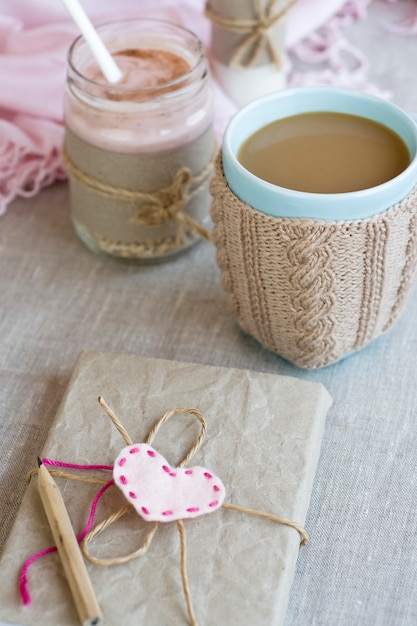 Avena, con sabor a yogur, espolvoreado con canela en un frasco de vidrio. Lo siguiente es una taza de café y una botella de leche. El siguiente es un cuaderno de manualidades y una servilleta rosa.