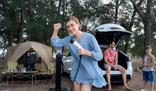 Aventura al aire libre y vacaciones familiares acampando en el mar viajes en automóvil ecológico Mujer alegre o madre sosteniendo el punto de carga EV con postura juguetona y feliz en el campamento Perpetuo