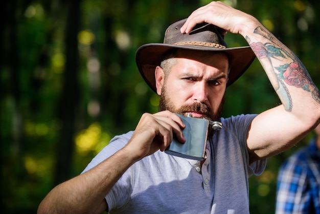 La aventura comienza desde el primer sorbo Bebida alcohólica Hombre nómada lleva alcohol con él Chico brutal vaquero barbudo bebe alcohol matraz de metal Hipster con barba bebe alcohol naturaleza fondo desenfocado