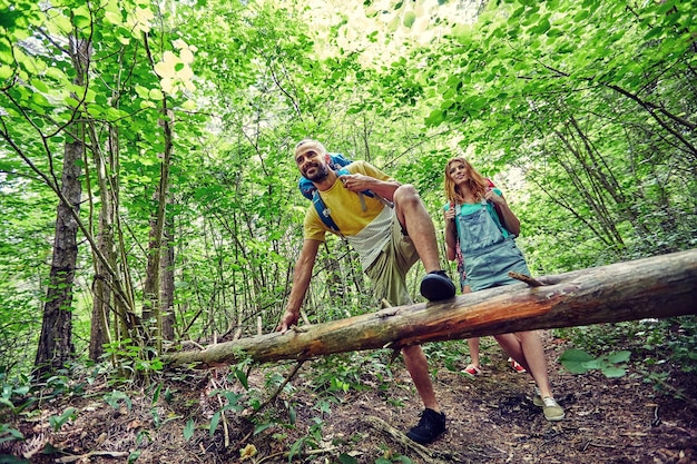 aventura, viagens, turismo, caminhada e conceito de pessoas - grupo de amigos sorridentes andando com mochilas e escalando tronco de árvore caído na floresta