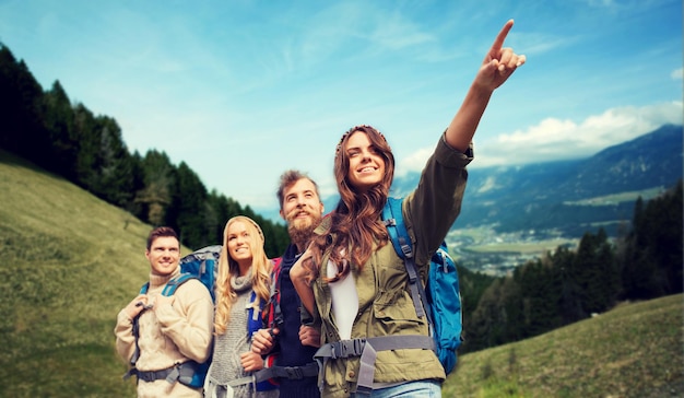 aventura, viagens, turismo, caminhada e conceito de pessoas - grupo de amigos sorridentes com mochilas apontando o dedo sobre o fundo das colinas alpinas