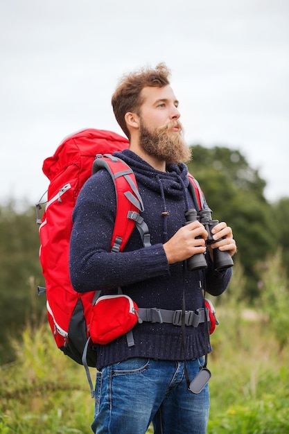 Foto aventura, viagens, turismo, caminhada e conceito de pessoas - homem com mochila vermelha e binóculo ao ar livre