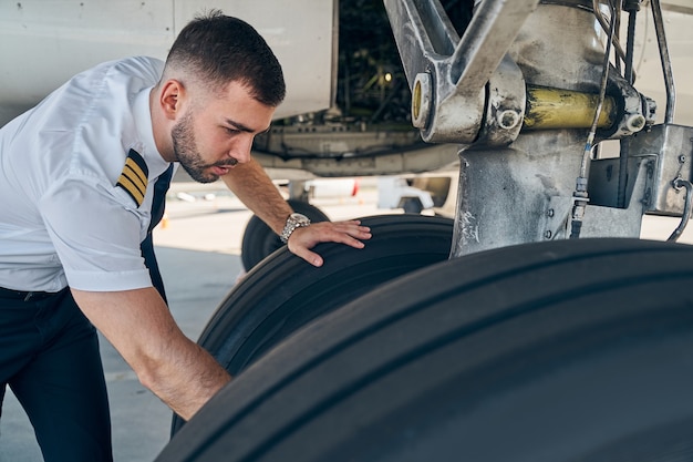 Aviador de cabelo curto focado e qualificado em uniforme verificando o sistema do trem de pouso do avião antes do voo