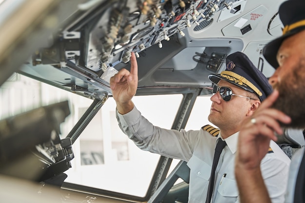 Aviador experimentado tranquilo con gafas de sol negras presionando los botones en el tablero mientras trabaja en una cabina con un colega