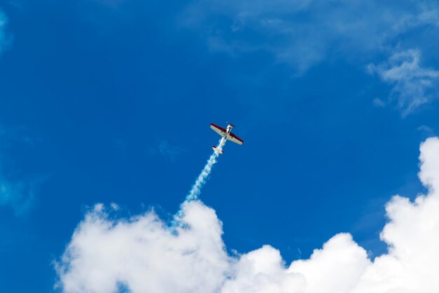 Avião esportivo vermelho e branco voa liberando um fluxo de fumaça no céu azul com nuvens