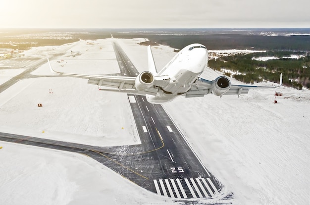 El avión es una vista alta del nivel de vuelo de subida en el aire, el aeropuerto de invierno de la pista, ciudad, nieve, bosques y carreteras.