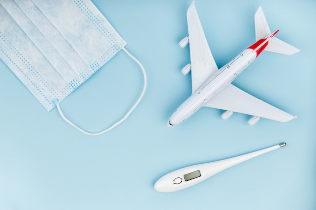 Foto avión, máscara médica y termómetro sobre un fondo azul.