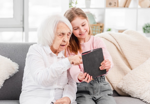 Avó com a neta sentada no sofá, olhando para a tela do tablet e sorrindo