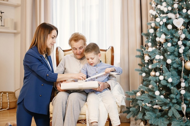 Avó com sua filha e neto no dia de Natal. Mulheres e garotinho abrindo um presente. Mulheres e menino vestindo roupas bege e azuis.