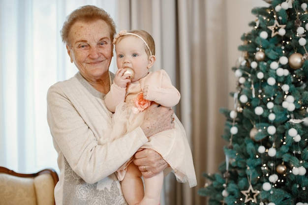 Avó com sua neta no dia de Natal. Velha segurando a menina em pé perto da árvore de Natal. Mulher e menina vestindo roupas bege.