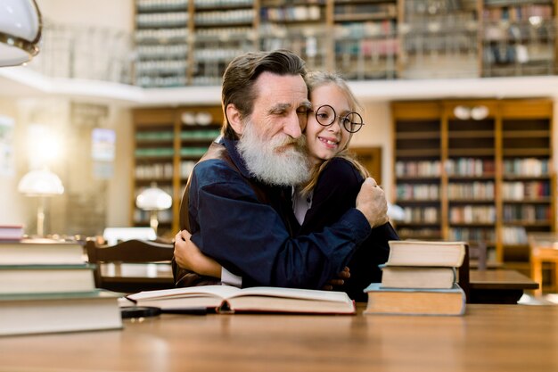 Avô e neta ou professor senoir e aluno aluno, sentado à mesa e abraçando-se, na antiga biblioteca da cidade vintage. Leitura, conceito de educação