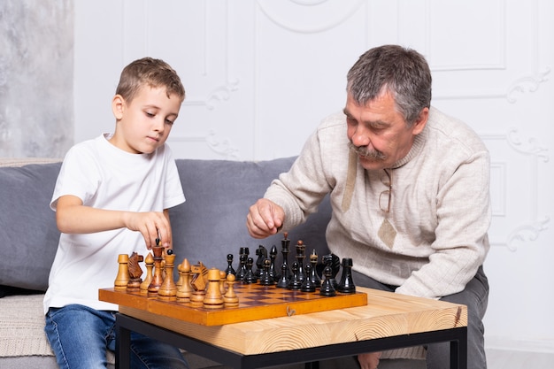 Avô jogando xadrez velho com neto interno. o menino e seu avô estão sentados no sofá da sala e brincando. último homem ensina uma criança a jogar xadrez