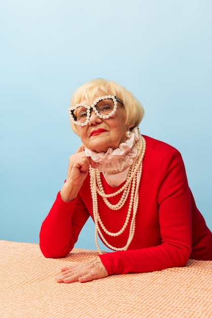 Avó linda velha em elegante vestido vermelho colar de pérolas e óculos posando sobre fundo de estúdio azul Conceito de moda de idade e emoções