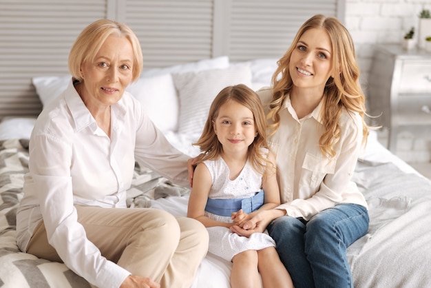 Avó, mãe e sua filha pequena sentadas na cama e coladas uma à outra, enquanto as duas mulheres abraçam uma criança e sorriem.