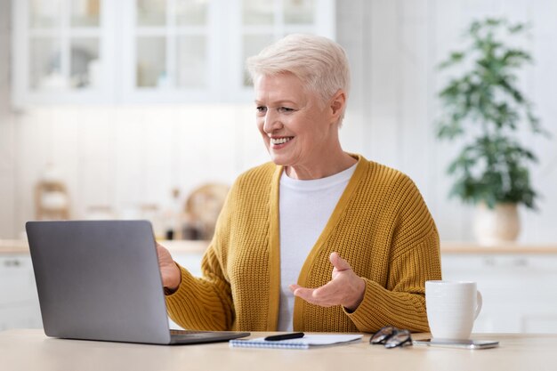 Avó positiva fazendo videoconferência com a família, usando um laptop moderno enquanto bebe chá na cozinha em casa, olhando para a tela do notebook, sorrindo e gesticulando, copie o espaço