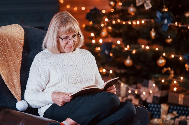 Avó sentada na cadeira com o livro na sala festiva de ano novo com decorações de Natal.