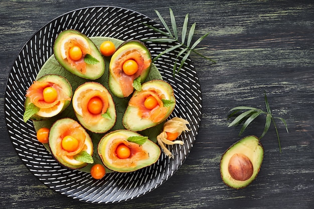 Avocado-Boote mit geräuchertem Lachs, Zitrone, Physalis und Minzblatt, Draufsicht auf dunkle Wand